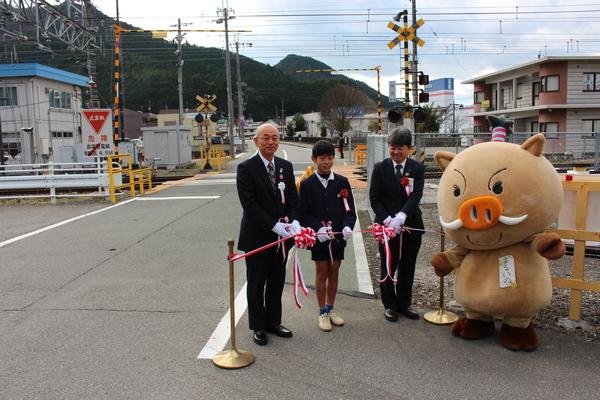 制服を着た小学生男子が市長らと一緒に弁天踏切の手前でテープカットしようとしているのを横で見ているゆるキャラの写真