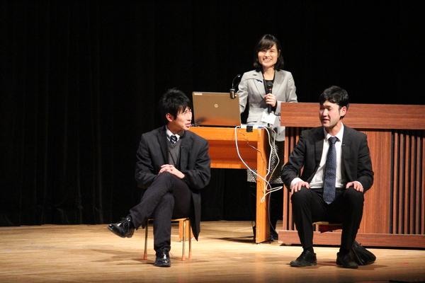 男性2人が小さい椅子に座って話をしていて後ろで女性が立って微笑んでる写真