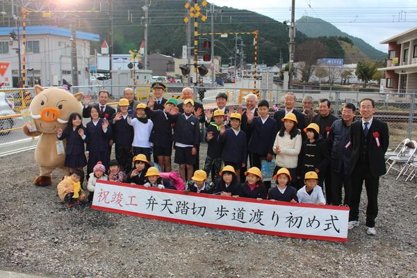 弁天踏切の手前の広場で「弁天踏切歩道渡り初め式」のパネルを小学生たちが持ち、JR福知山支社の前田支社長や市長らと一緒に記念撮影している写真