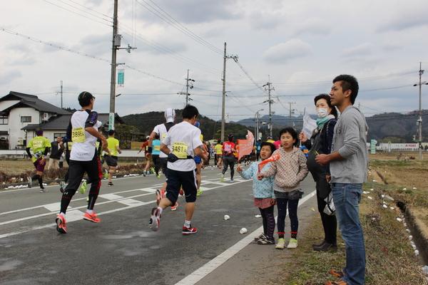 沿道で家族連れで応援に来ていて、お母さんはマスクをして、子供達は旗を持って応援している写真