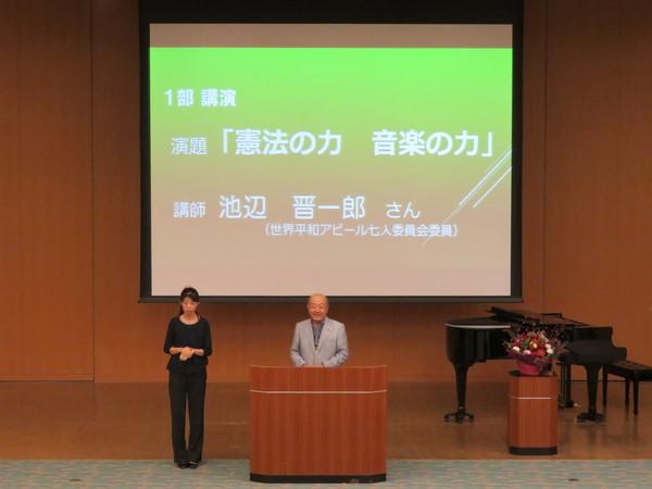 1部講演演題「憲法の力音楽の力」講師池辺 晋一郎さん(世界平和アピール七人委員会委員)と書かれたスクリーンの前に立っている池辺 晋一郎さんとその横に立っている女性の写真