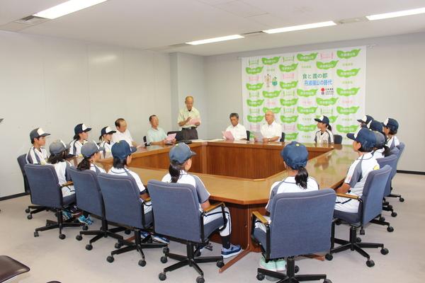 市長と、関係者と、少女ソフトボールチームのラクーンズ・ジュニアのメンバーがユニフォームをきて、会議テーブルに着席している写真