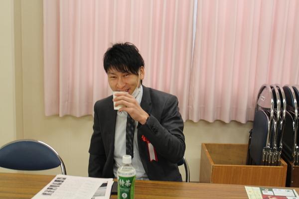 控室で胸に赤いリボンを付けた男性が紙コップでお茶を飲む写真