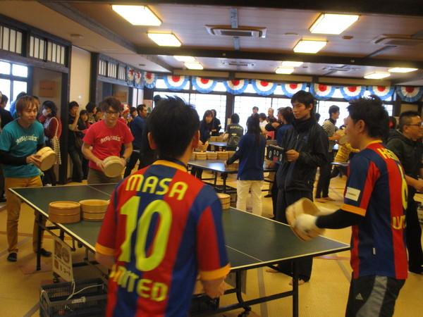 卓球台のネットの変わりに桶が中央に置かれており、会場に沢山の卓球台が設置され、2人1組のペアで、それぞれの試合が行わている様子の写真
