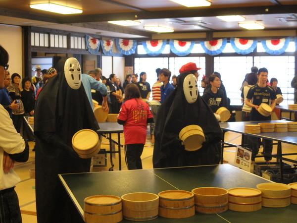 映画のキャラクター「かおなし」の仮装をした二人が桶を手に持って卓球台の前に立っている写真