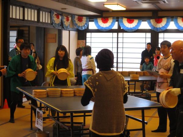 緑の法被を着た男性と黄色の法被を着た女性が桶を持って卓球台の前で構えており、相手チームの茶色いセーターを着た女性の後姿が中央に写っている写真