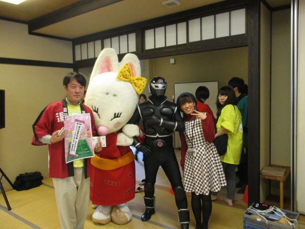 赤い法被を着た男性と、チェックのワンピースを着た女性がきつねの着ぐるみの人形と 霧海の戦士 ササグローと一緒に写っている写真