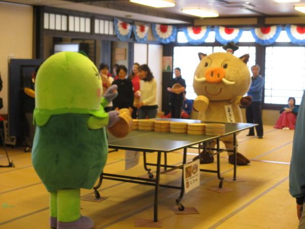 まめりんとまるいのが試合で対戦しょうと、卓球台の前で桶を持って向い合っており、まめりんがボールを持って打とうとしている様子の写真