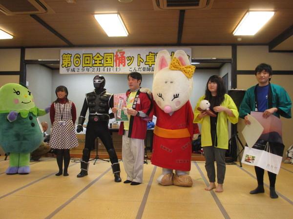 第6回全国桶ット卓球台大会の横断幕の前に、左からまめりん、チェックのワンピースの女性、ササグロー、赤い法被を着た男性、きつねの着ぐるみを着た人形、黄緑色の法被を着た女性、緑色の法被を着た男性が写っている写真