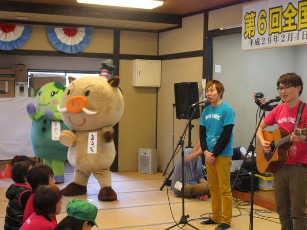 ギターを持った男性がギターを弾きながら、と青いティーシャツを着た男性と、スタンドマイクの前で歌を歌っており、その左隣りで、桶を持っているまるいのとまめりんが音楽に合わせて踊っており、その様子を大会参加者が畳に座って見ている様子の写真