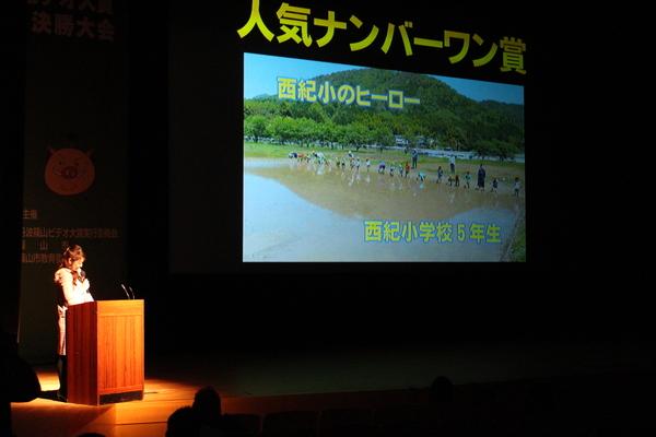 「人気ナンバーワン賞」を受賞した西紀小学校の映像が舞台のスクリーンに映し出されており、演題で女性司会者が紹介している写真