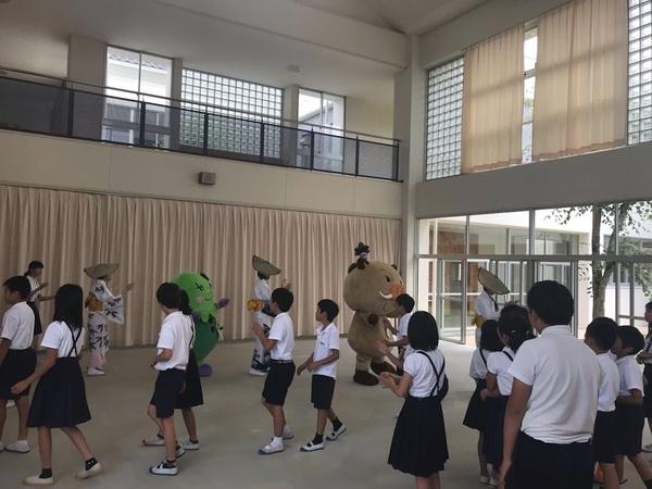 マスコット人形とおけさ傘を頭に被った白色の着物姿の踊り手3名に加わり見様見真似で生徒全員も手拍子をしながら踊りをしている写真