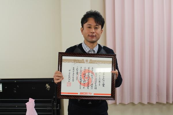 男性が額に入った賞状を持って記念撮影している写真