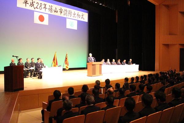 出初式で市長が挨拶しているのを壇上で聞いている人々と客席で聞いている人々の写真