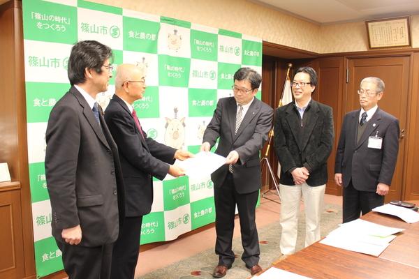 篠山市商工会、丹波篠山観光協会、丹波ささやま農協の関係者が市長室を訪れて、市長が隣のメガネをかけて茶色の革靴の男性より要望書を受け取っている様子の写真