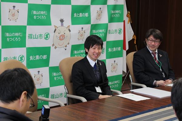 羽馬 雅人さんが笑顔で椅子に座っておりその隣に関係者が座っている写真