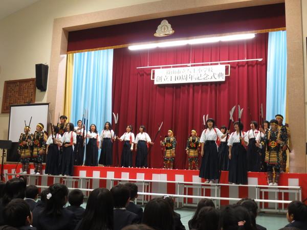 創立140周年記念式典と書かれている横断幕のある舞台の上で、袴を着て槍を持っている女子児童と甲冑を身にまとい槍を持っている男子児童が舞台の上に立って発表している写真