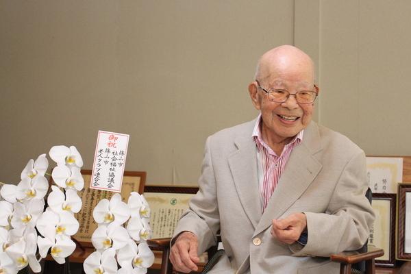 白い欄の花の横に座っている波部 時雄さんの笑顔の写真