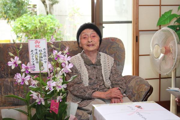 ご高齢の女性がソファに座り、周りには、お祝いの花等がある写真