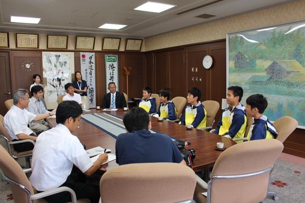 市長室にて、西紀スイミングスクールの生徒5名が黄色と青色の入ったジャンパーを着て座っており、前方に座った教育長の話を聞いている様子の写真