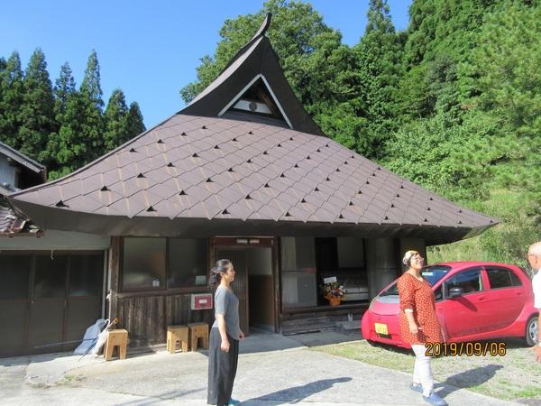 特徴的な屋根の家屋の前の玄関前に立つ2人の女性の写真