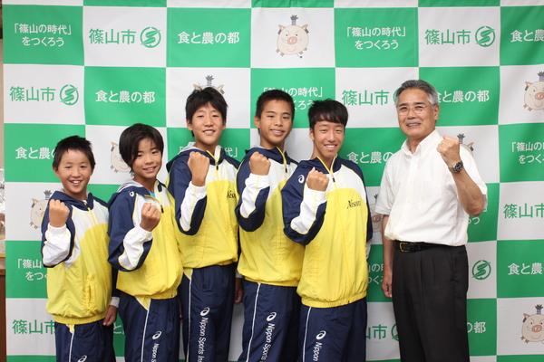 西紀スイミングスクールの生徒5名が黄色と青色の入ったジャンパーを着て、教育長とガッツポーズをして写っている写真