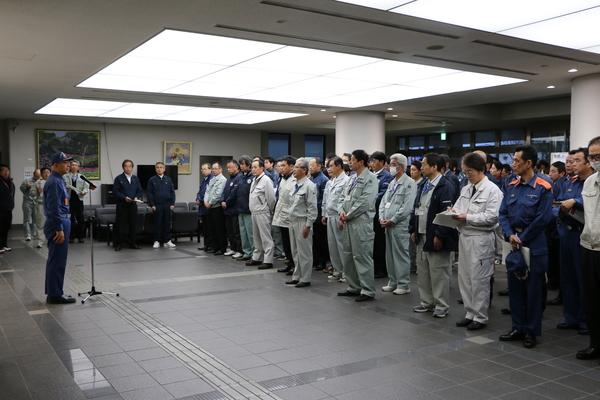 ロビーにて消防の帽子を被り消防服姿の市長がスタンドマイクで話しているのを整列している作業服姿の職員らが聞いている写真