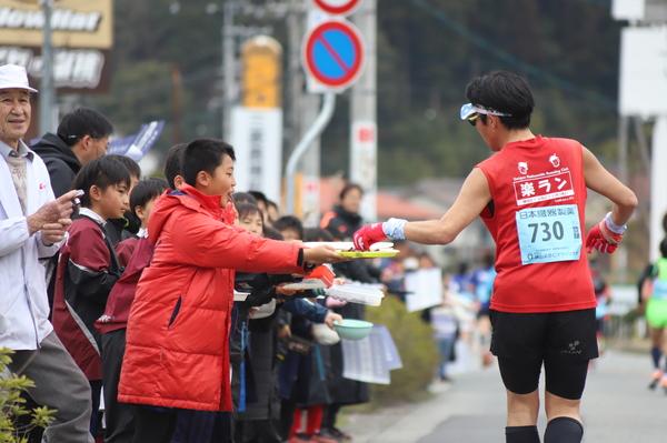 赤色のTシャツと帽子にサングラスをかけたランナーに沿道でお皿にのった食べ物を差し出している子供の写真