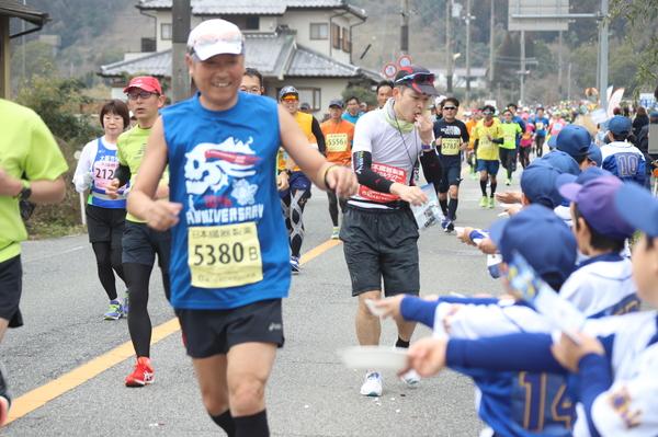 走ってくるランナーに沿道からお皿に入った食べ物を差し出しているユニフォームと帽子を被った子供たちの写真