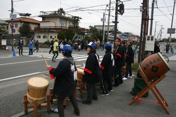 沿道脇で頭にハチマキをした男性や帽子を被った女性が太鼓を叩いて応援している写真