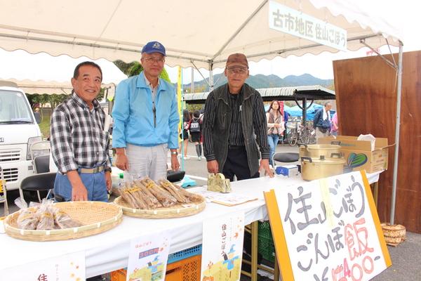 テントの中で販売用のごぼうとごぼう飯を前に男性3名が写っている写真