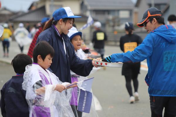青色のウインドブレーカーを着たランナーにお皿に入った食べ物を差し出している女性の写真