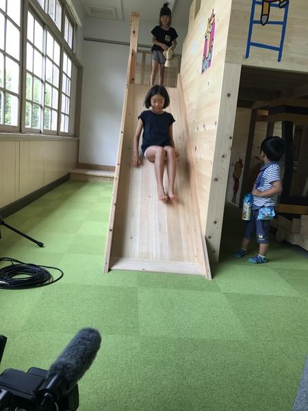 木で作られた滑り台を女の子が滑っていて、後ろで他の女の子が滑ろうと待っている写真
