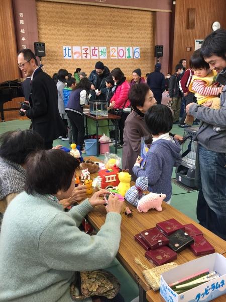 体育館の中の長机でそれぞれ分かれて作品が紹介され、子供や大人がその作品を見ている写真