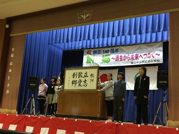 校訓「立志、敬愛、創郷」と書かれた大きな額を子供達が舞台から紹介している写真