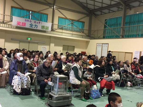 体育館には業務用のストーブがたかれ、お客さんたちがお話しを聞いている写真