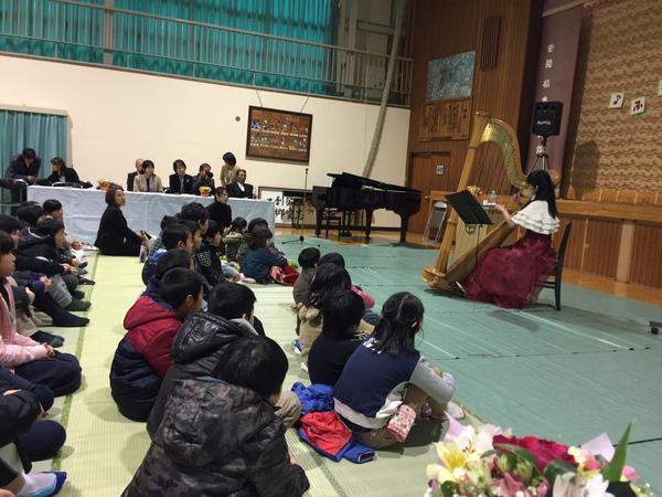 女性が椅子に座りハープを弾いてその音色を子供達が静かに座って聞いている写真