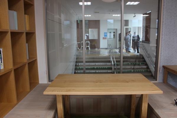手前には木のテーブルがあり、その後ろに透明の窓ガラスがあって、奥にいる人たちや階段が見えている写真
