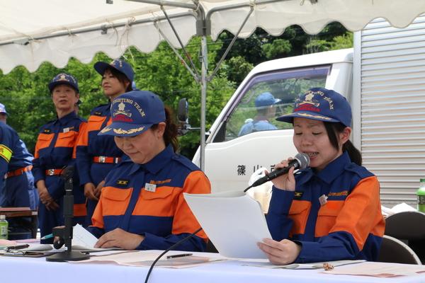 帽子を着用した作業服姿の女性消防団員が2名テントに座っており一人がマイクでアナウンスをしている写真