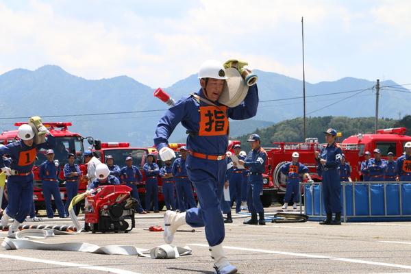 ヘルメットを着用している作業服姿の消防団員が消防用ホースを担いで走っている写真