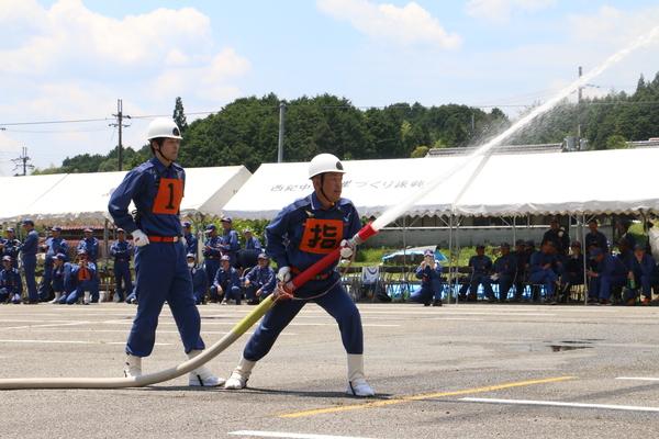 ヘルメットを着用している作業服姿の消防団員がホースから放水している様子をもう一人の消防団員が見ている写真