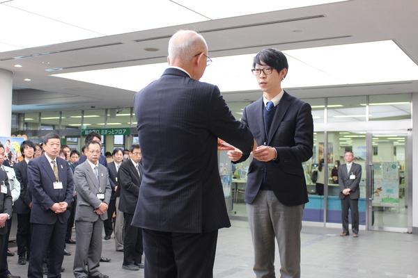 職員の前で、市長が中森恵佑君へ書類を渡している写真