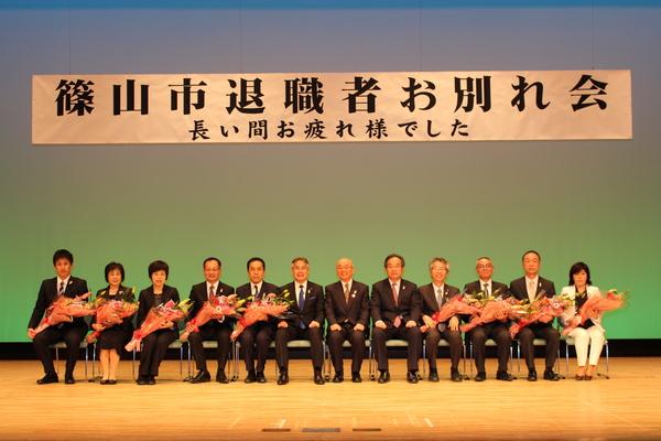 市長を中央に9名の花束を持った退職者達が椅子に座り記念撮影をしている写真