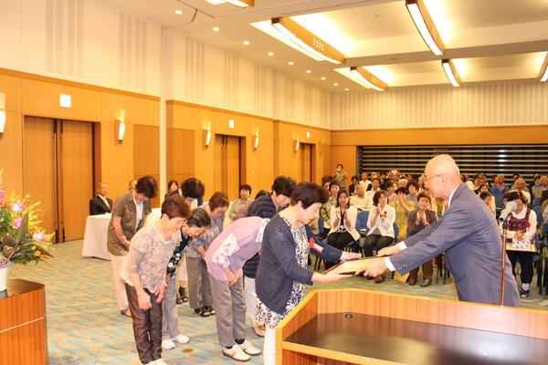 8人の女性グループの代表者1人に賞状を手渡し、他のメンバーはお辞儀をしている写真