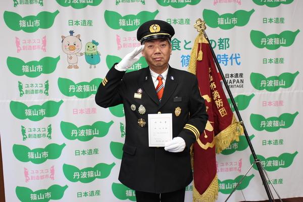 消防団の旗の横で、敬礼し、手に白い紙を持つ制服姿の澤 光吉さんの写真