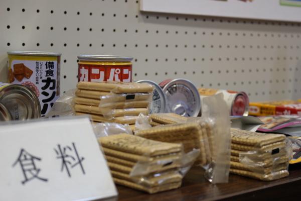 机の上に「食料」と書かれたネームプレートが置かれており、カンパンやクッキーなどの非常食が並べられている様子の写真