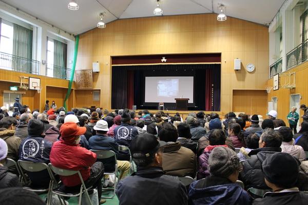 体育館に沢山の住民が集まって椅子に座っており、舞台の上のスクリーンの映像をみている様子の写真