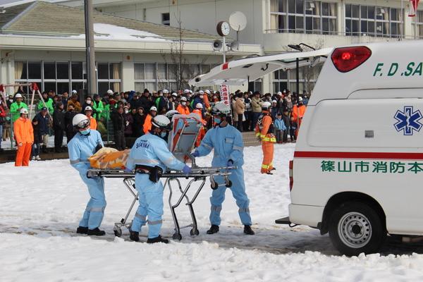西紀南小学校の運動場に救急車が泊まっており、担架に乗せて救急隊員が救急車に乗せようとしている様子を後方で住民が見ている様子の写真