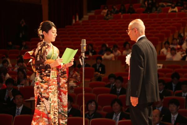 壇上にて振袖を着た女性が、市長の前で、黄緑色の紙にを手に持って文章を読んでいる様子の写真
