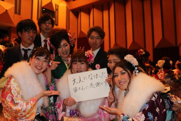 「大人になりました。皆さんに感謝です。」と書いた画用紙を持って写っている振袖を着た新成人5名とその後ろにスーツを着た男性3名が写っている写真